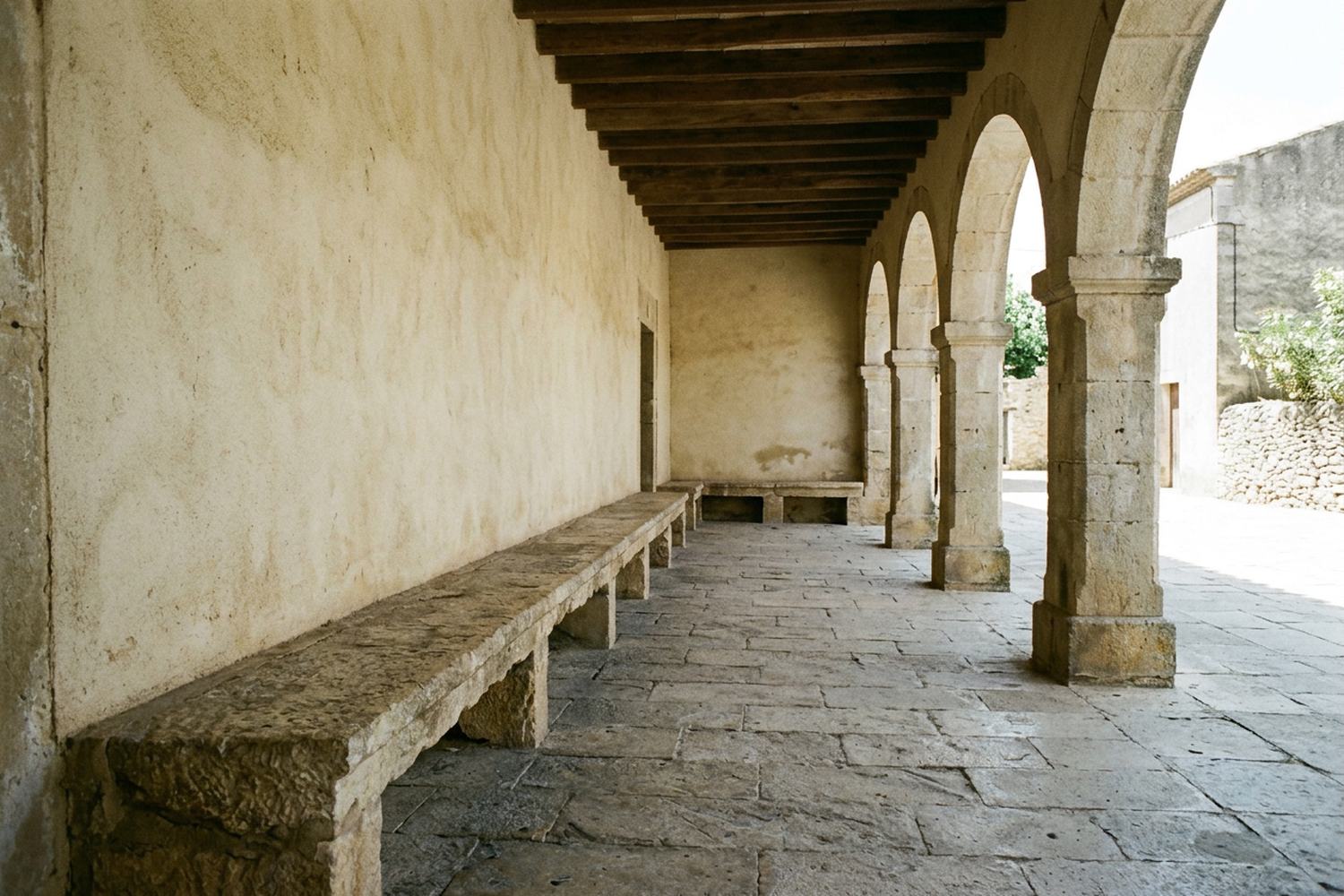 A long stone bench in the shade of an arcade beside sunlit paving.
